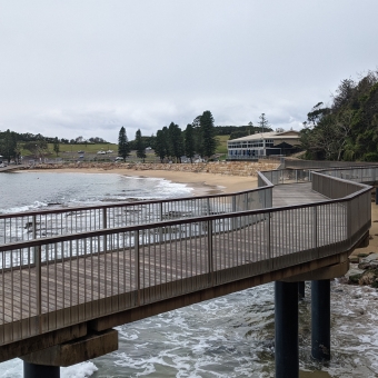 Walkway Overlooking Terrigal Beach, with grey sky at the top, reflected in grey, calm-ish surf to the left and bottom, with a thin stretch of land in the centre stretching towards rocks on the right of the image, with a walkway seemingly hovering from bottom left to middle right of image.