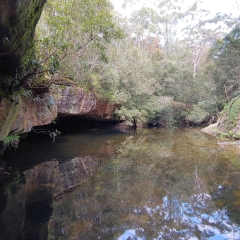 Berowra Creek Under Historic Military Steele Bridge; left picture rocky outcrop overhanging water, with trees on top overhanging further, bottom and middle covered in wide expanse of water going to the back and right, trees lining the creek into distance.