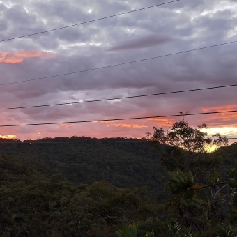 Sunset From Bimbil Track; bottom has silhouetted hill with gum trees, top has rolling clouds with dots of sky through and streaks of horizontal red spreading from just behind the hill after sunset.