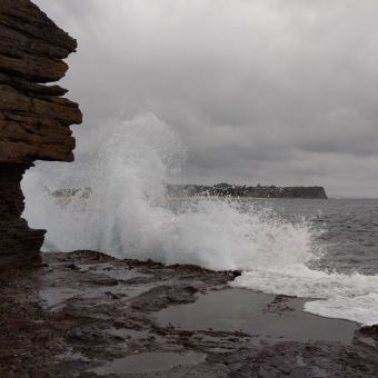 Headland Splash, with rocky cliffs on the left, grey wrinkly ocean on the right, rock flat with pooling below, and grey storn clouds above, with a white splash of water far in front of Mona Vale Beach in the distance