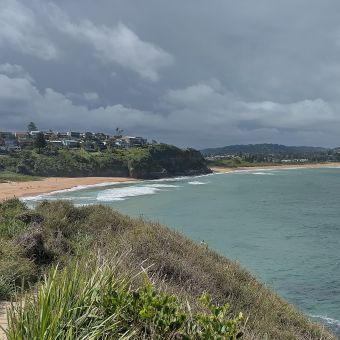 Warriewood and Mona Vale Beaches, with Turimetta Headland covered in ferns in the bottom left corner, the beaches going off into the distance in the middle, and storm cloulds rolling over the top of the picture, with turquoise flat ocean on the right of the picture