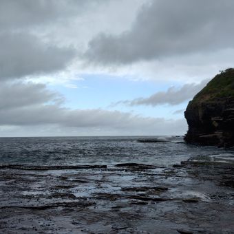 Blue Patch Of Cloud, with grey storm clouds parting to reveal a light blue hole of sky, with flat grey ocean below and rocky cliffs covered in green shrubbery to the right