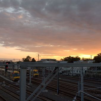 Sunset Above The Trainyards, Hornsby, with grey, fluffy clouds lined with streaks of gold eminating from the centre right, with two parked trains in the bottom left corner on tracks which lead to the bottom right corner