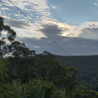 The Clouds' Sunset, with dark green trees fading away into the valley into much darker trees on the other side, with the sun setting behind grey, slightly streaky clouds which gives them a golden edge