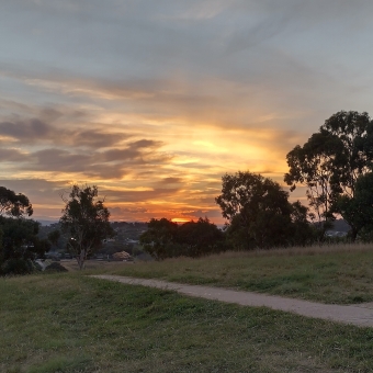 Phosphorescing Sunset, Hill Reserve, Ngunnawal, with a footpath cutting across the foreground, disappearing over a grassy hill to the left, with silhouetted full-grown gum trees smalled by the glowing clouds dominating the top of the frame, striped with orange, grey, and faded blue sky, with a lava-like sun barely visible on the horizon