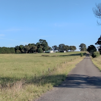 The Lone Farmstead From Darling Lane, Exeter, with a straight paved road on the right disappearing over the horizon, with a open, green, grassy field on the left only surrounded by a low wire fence which leads up to a distant white house with a green roof which is surrounded by what are large trees but look small because they are so far away