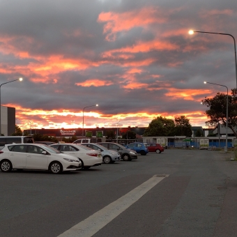A Fire-Like Sunset, Gungahlin, with a red hot glow from the setting sun on the horizon barely visible above a shopping centre, with a carpark filled with streetlights and different-coloured cars in the foreground