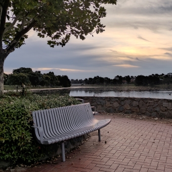The Soroptomist's Seat, with a metallic seat in the foreground backed by a low hedge and single elegant tree, all of which is surrounded by a bricked walkway and low stone wall, with the seat looking out over the lake and silky clouds transitioning from grey through peach to golden