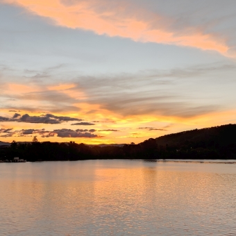 Sunset at Lake Burley Griffin, with cloudy streaks of orange, grey, and baby blue through the sky reflected in chequered waves of the lake, with Telstra Tower atop Galambary on the right and the National Museaum on the left silhouetted against the golden sky