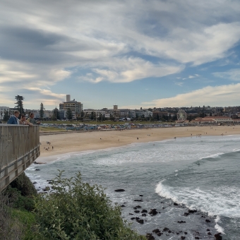 Typical Australia, Bondi Beach, Bondi, with wispy clouds almost covereing a barely discernible blue sky, with rolling waves on the right reflecting grey and chequered with white, with a sandy beach dotted with people and a strip of mixed buildings hiding the horizon, with a balcony on the left with six people spread out along it and all of whom are looking out over the previously described expanse