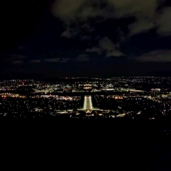 Canberra Awake As Ever, with the centre of Canberra outlined with the tiny dots of light which are whole buildings and street lamps, with ANZAC Parade lit up at the very centre of the picture and Parliament House just above it, with darkened clouds in the sky barely reflecting the light of the city