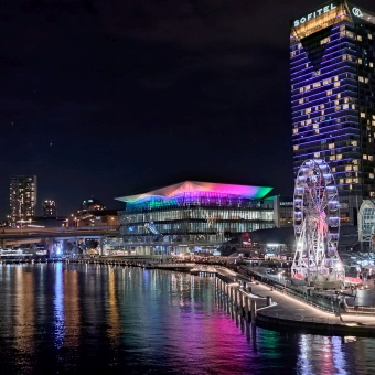Colourful Darling Harbour, Pyrmont Bridge, Sydney, with Darling Harbour Theatre at the centre of the picture with its roof lit up in varied neon colours, with the Sofitel skyscraper standing tall to the theatre's right which is coloured navy blue with some gold-yellow windows and trim near the top, with the reflections of these lights and other background city skyscrapers in the waters of Darling Harbour below, with the night's sky the darkest navy blue which one only finds lit up by the city ambience