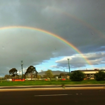 Double Rainbow From The Tram, near Dickson, with two semi-circles of rainbow dominating the grey sky, with a road, a couple of trees and light poles dwarfed at the bottom of the image