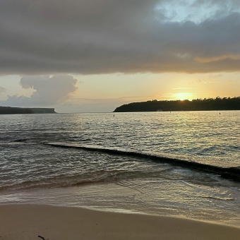 Balmoral Beach, Mosman, with North Head and Middle Head covering the horizon except for the middle fo the picture which the ocean extends as far as the eye can see, with the sun rising over Middle Head and reflecting off both the flat clouds above and the ripplings water below, with the visible sky turning from golden to baby blue