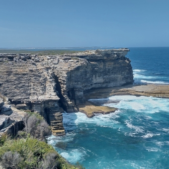 The Balconies, Royal National Park, with a large rocky outcrop extending into the bluest of blue oceans from left to right, with two fisherman dwarfed by the magnitude of the outcrop