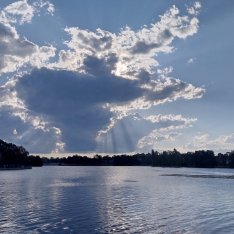 Sun Rays, Yerrabi Pond, Gungahlin, with a singular triangular grey cloud in the centre of the sky and the sun shining its rays around the cloud like a hug, with rippling water reflecting the grey and blue of the sky, with water and sky divided by a dense line of silhouetted trees