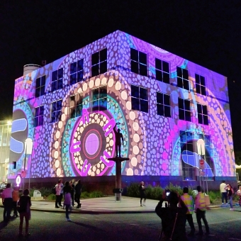 Questacon, with people silhouetted in front of it, with the exterior lit up with Indigenous dot paintings representing a campsite and a water way, with mostly purple dots but with some circles of yellow dots and some of teal dots