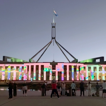 New Parliament House, with both wings half visible, with the pylons at the front lit up with a multitude of colours in homage to the LGBTQ flag, with about fifteen people in a line silhouetted with their high-tech cameras