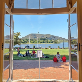 Eating Room View, with an opened door frame looking out over a meticulously mown lawn dotted with picnicking people, with four flagpoles at the other end of the lawn which contain the Australian and Aboriginal and Torres Strait Islander and Australian Capital Territory flags, with Lake Burley Griffin then Black Mountain extending off into the distance, with the latter having Telstra Tower almost centre frame atop it