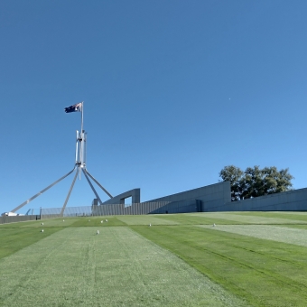The Lawns of Parliament, Parliament House, with the green grassy slope of one of the sides of the House extending from the bottom to about half way up, with grey cement-brick walls dividing the grass with the bluest sky, with the spire and the Australian flag slightly to the left of centre, but extending far up into the sky
