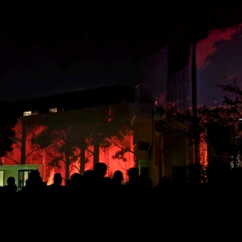 Portrait Gallery Ablaze, with a deep red bushfire amongst silhouetted trees prjected up to the entry point and left wing of the building, with silhouetted heaps of people inb front of the projection
