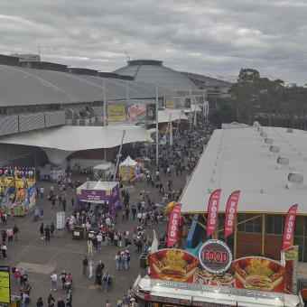 Crowded Everywhere, with the angle of the picture from above the crowds from in a ferris wheel, with hundreds of people in a large thoroughfare in the centre of the frame, with show pavillions to the left and right of ther crowds, with stalls in front selling various carnival foods, with grey clouds which still let through a good amount of light