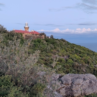 Lighthouse Over The Ocean, with the clean-cut sandstone Lighthouse on the left of the picture atop a green hill which slopes down towards the right, with the blue ocean appearing behind the hill as it slopes, with white fluffy clouds near the horizon in an otherwise blue sky