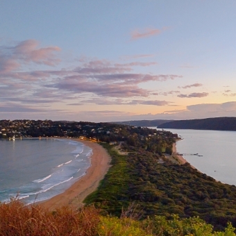 The Ocean-Bay Divide, with the sky above turning from baby blue at the top to peach-yellow at the horizon in the centre, with faded-purple clouds gilded in baby pink streaked across the sky, with a green peninsula stretching from the horizon to underneath the picture which has a pure-yellow beach lining each side, with think white waves crashing on the left beach