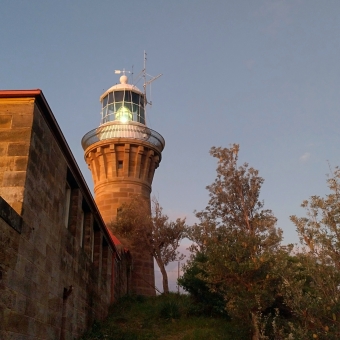 A Long Night Ahead, with the sandtsone Lighthouse standing tall slightly to the left of centre, with the large light visibly lit on the inside, with a sandtsone wall on the left of the main lighthouse, with seemingly fluffy khaki-green trees on the right, with a clear blue sky in the background and the top half of the picture