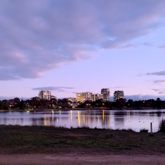 Purple Haze, Yerrabi Pond, Gungahlin, with silhouetted trees left and right and a dirt track below in the foreground, with the sky which turns from pink to baby blue streaked with purple clouds which are gilded with an even brighter pink, with the horizon covered in half a dozen how high-rise buildings with all their yellow windows illuminated, with the lake between the dirt path and horizon reflecting the yellowed buildings and purpled sky