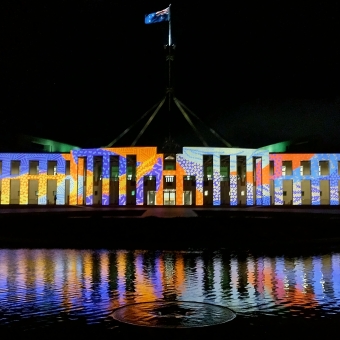 Reconciliation Lights, New Parliament House, with New Parliament House from and centre seemingly covered in Indigenous art which is a mosaic of coloured pattertned dots and dashes which has some sections of blue and some of yellow and some of orange and one of purple and one of pink and one of green, with the Australian flag visible atop its mast, with the sky behind pitch black, with the reflection of all light in the shallow pool of water below