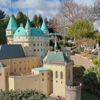 The Slovakian Bojnice Castle, with a mimature replica of the castle on the left and part of Cockington Green Gardens off into the distance on the right, with beige cement-rendered walls on the grandiose castle which also has teal roofing, with many bonsai trees in the surrounding gardens