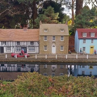 Canal Townhouses, with three different buildings abutted agains each other which are a Tudor house and a grey brick house and a blue wood house, with a straight road with a red convertible and human figurines on it with a dark blue canal in the immediate foreground