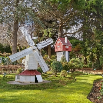 The Windmill and Its Farmhouse, with a white windmill in the foreground on the left which has perfectly manicured grass and bonsai trees around it as well as a redwood house on a highrise behind it in centre frame, with low largens immediately around these structures and a larger garden with full size trees surrounding the image