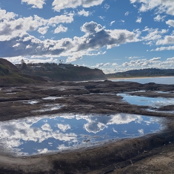 Turimetta Head, Warriewood, with blue and white chequered sky reflected in a clear and circular rook pool which is front and centre, with the rest of the rocky cliff face appearing small by comparison, with distant beaches and coastlin in the far distance