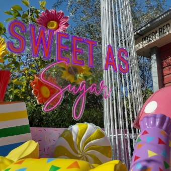 Sweet As Sugar, The Grounds Of Alexandria, Alexandria, with flourescent and multicoloured large plastic ornaments shaped in various lollies throughout the bottom of the image, with the words 'Sweet As Sugar' in neon lighting above and just to the left of centre, with clear blue sky, a green shrub, and the corner of a red-brick building filling the background