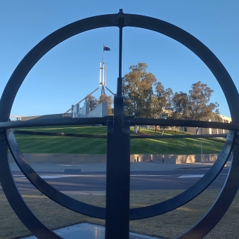 Parliament House, Canberra, with a metal spherical artwork in the immediate foreground which divides the image up into equal quarters, with the spire of Parliament House in the top left quarter, with gum trees in the top right quarter, and with the sloping grassy hill and road stretching between the two bottom corners