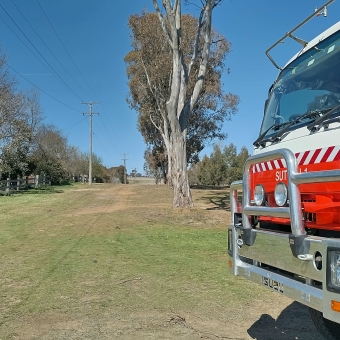 Bob Collis Reserve, Sutton, with the front of a Rural Fire Service truck in the immediate foreground on the right of the image, with a line of trees mirroring telegraph poles leading into the distance in the centre of the image, with clear blue sky in the background