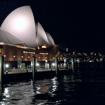 Sydney Opera House, Bennelong Point, with the curved sails of the Opera House in the foreground on the left of the image, with dots of lights from distant buildings on the right of the picture, with reflections of all these in the seemingly black water, with an equally as black sky
