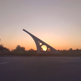 Giant Sundial, Singleton, with a five-metre tall sundail in the middle distance which has a circular hole in it which perfectly frames the sun setting on the horizon, with silhouetted trees dotting the horizon, with an orange glow emanating from the horizon which turns blue-grey the further away from the horizon it stretches
