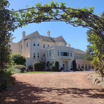 Government House, Yarralumla, with the eastern side of the Govenor General's residence in the centre of the picture, with a wide orange gravel path leading up to the side steps, with a treselled hedge framing the other three sides which is in front of the clearest blue sky