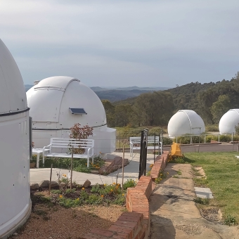 Mt Stromlo Observatory, Weston Creek, with two dome-topped telescopes in the left-hand immediate foreground and three in the right-hand middle distance, with a much larger dome just visible in the far distance on the right, with a paved path joining the five closer telescopes and the rest of the foreground covered in grass, with with every undulating hill towards the horizon thickly covered in dark green trees