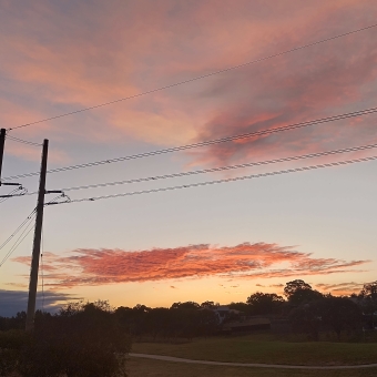 Rich Sunset, Gungahlin Lakes Golf Course, Ngunnawal, with tall and buzzing powerlines cutting through the sky which is streaked with brilliant orange clouds on an otherwise pastel blue sky which slowly changes to pastel orange as it gets closer to the horizon, with the barely visible green of the gold course below