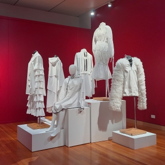 White-Out by CIT Fashion School, Canberra Museum & Gallery, Civic, with six stands which have various types of white clothing hanging on display in the middle of a small-to-medium room with red walls