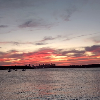 Silhouetted City, Watsons Bay, with clouds streaked near the horizon glowing red and glowing orange in an otherwise pastel blue sky, with Sydney City skyline silhouetted just on the horizon which has Centrepoint Tower towards the left and Barangaroo towards the centre and the Harbour Bridge towards the right as distinctive silhouettes, with the rippling water below reflecting the colours of the sky above and occassionally dooted with snall personal yachts