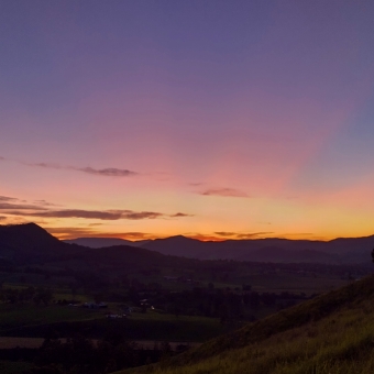 Sunset from Kia Ora Lookout, Gloucester, with otherwise green valley and mountains cast into shadows, with a deep orange glow at the horizon turning into blue at the top of the picture, with peach-coloured rays emanating from the sun and the tops of mountains cutting shadows through them