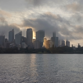 Sydney CBD from Mrs Macquaries Chair, Sydney, with highrise on centre frame with a singular golden beam of light from the sun peering through in the direct middle of the frame, with storm clouds above, with grey and rippling water below