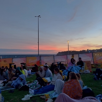 Sunset Cinema, Bondi Beach, with the horizon over the water streaked with pastel pink and puruple and peach, with people sitting on picnic blankets in the foreground facing to the left, with baby blue sky above streaked with barely visible grey clouds