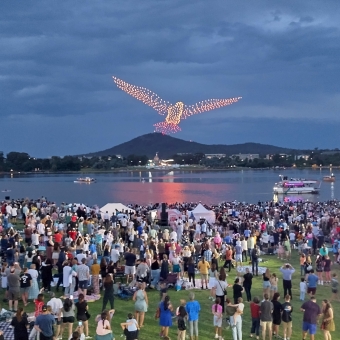 Gamburra Totem, Flight Drone Show, Commonwealth Place, with about six-hundred drones appearing as bright dots of light arranged into a bird and hovering over a lake, with many people standing on the low and grassy slope in the foreground, with grey-blue clouds densely packed above