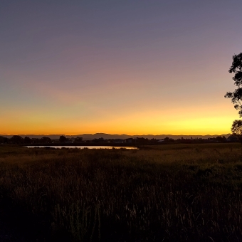 Radiating Sunset, Little Mulligans, Bonner, with a glowing orange sun strewn across a cloudless sky changing from gold to orange to magenta and finally fading to blue, with grass turning into low rolling hills silhouetted below